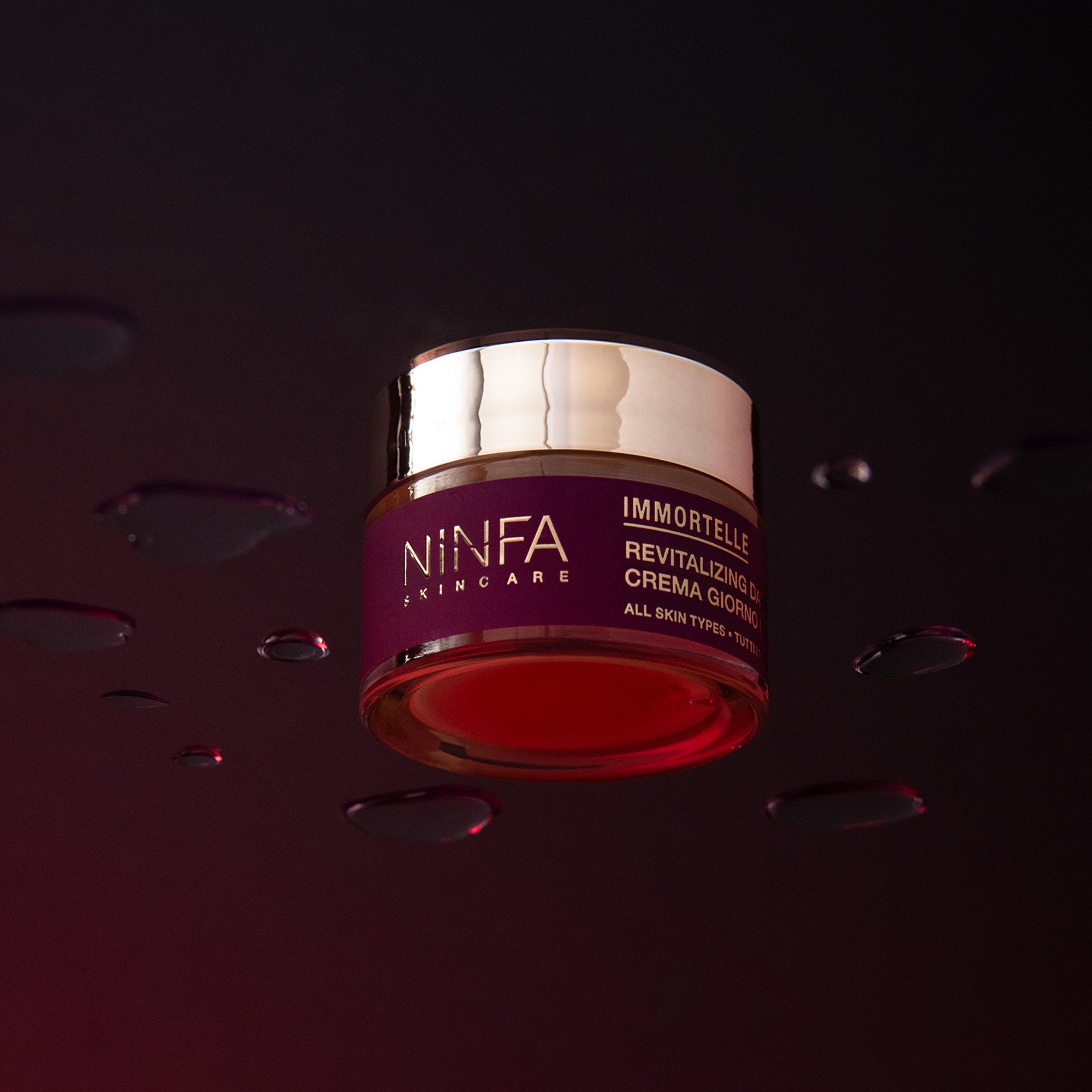 Primo piano della crema viso rivitalizzante all’elicriso di Ninfa Skincare su vetro con gocce e sfondo viola scuro
